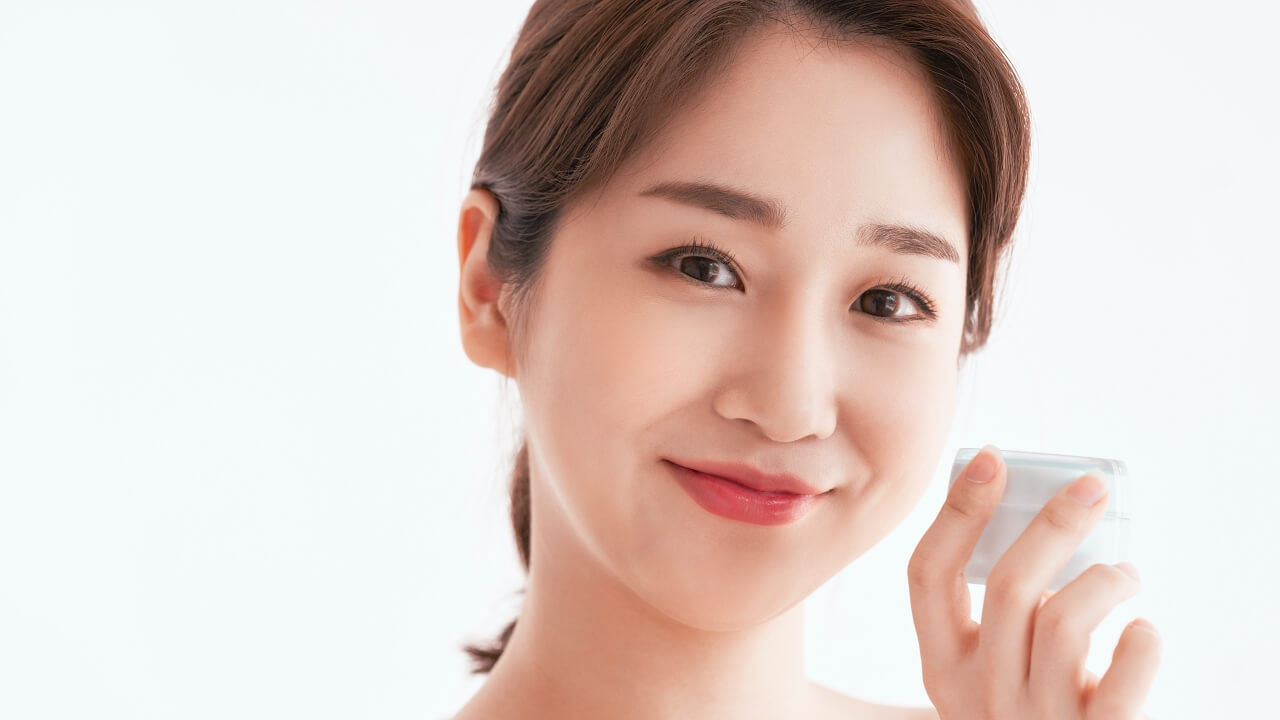 一位年輕的亞洲女性剛做完皮秒雷射，對著鏡頭溫柔地微笑，臉頰附近拿著一小罐精華液，背景是明亮的白色。